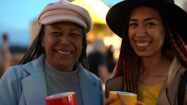 Happy african mother and daughter having fun together drinking hot drink at christmas street market druing winter time - Focus change from faces to drinking cup
