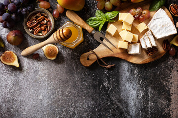 Brie cheese, fresh figs, grapes, cheese cubes, pears, pecan nuts and honey on cutting board on dark stone background. Cheese plate, still life of seasonal wine snacks. View from above. Copy space.