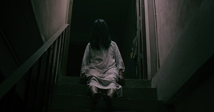 Horror scene of a mysterious Scary Asian ghost woman creepy have hair covering the face sitting on staircase at abandoned house with background dark scene movie at night, festival Halloween concept
