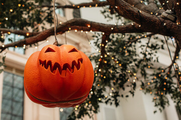 Scary pumpkin Halloween face hanging on a tree with twinkling golden garlands. Halloween party...