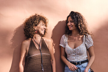 Mother and daughter posing smiling and in complicity in front of a brown wall.