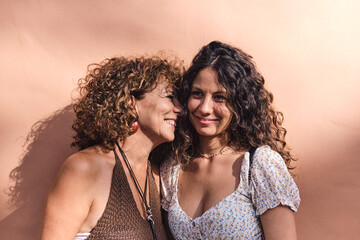 Mother and daughter posing smiling and affectionate in front of a brown wall.