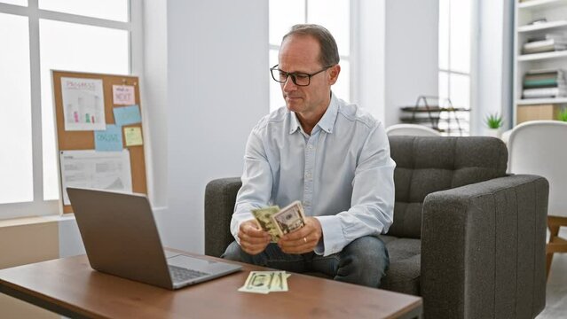Confident middle age man, a happy business worker, basking in his success as he sits in office interior, counting dollars with a smile on his laptop