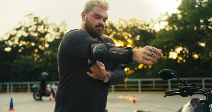 A man biker and strong build with a thick beard puts on elbow pads for protection while eating on a motorcycle