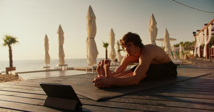 The guy does the stretch following the video on the tablet. Online stretching and yoga classes. Sunny beach in the morning