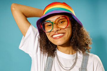 Smiling woman in rainbow cap and eyeglasses looks camera over blue studio background