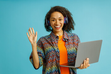 Smiling woman in headphones holding laptop and waving hi in camera on blue studio background