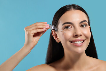 Beautiful young woman applying serum onto her face on light blue background