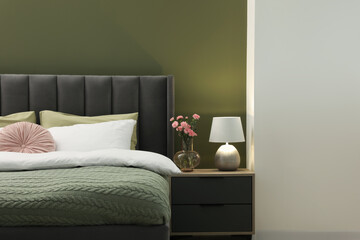 Large comfortable bed, nightstand, lamp and beautiful flowers in stylish room. Interior design