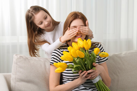 Daughter covering mother's eyes with her palms and congratulating with bouquet of yellow tulips at home