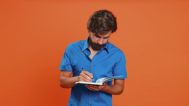 Thoughtful journalist young middle eastern man making notes, writing down thoughts with pen into notepad notebook diary, to do list, good idea inspiration. Arabian guy isolated on orange background