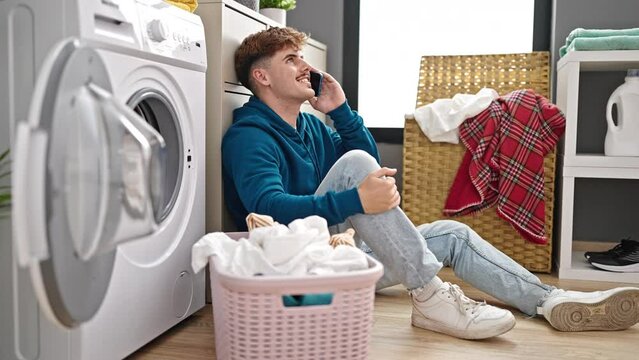 Young hispanic man talking on smartphone washing clothes at laundry room
