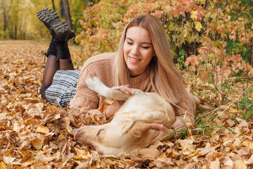 A young woman laying on the dry yellow leaves with her beloved labrador dog in autumn park.