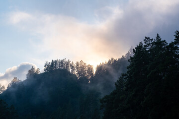 Mountains and peaks in fog at sunrise or sunset. Sun from behind the mountain. Amazing view of mountains and forest landscape with cloudy skies Altai mountains. Mountains and peaks in fog at sunrise