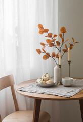 Autumn home interior in the living room - around wooden table, a chair, a bouquet of autumn leaves in a vase, decorative pumpkins. Cozy home