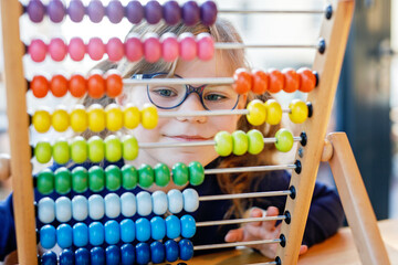 Little preschool girl playing with educational wooden rainbow toy counter abacus. Healthy happy child with glasses learning to count and colors, indoors on sunny day.