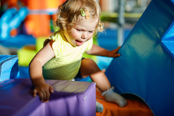 Happy blond little toddler girl having fun and sliding on indoor playground at daycare or nursery....