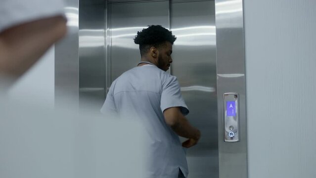 African American physician in uniform stands near lift door and presses call button. Medical worker waits elevator in medical centre corridor. Multiethnic healthcare specialists at work. Back view.