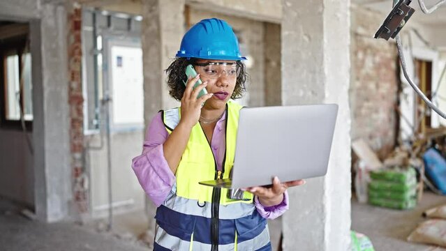Young beautiful latin woman builder talking on smartphone using laptop at construction site