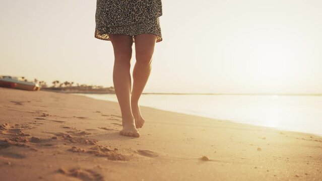 Barefooted woman in airy white dress walking on sandy ocean beach leaving footprints at sunset, rear view. Female, girl enjoying resting relaxing on vacation. Travel, tourism, nice evening concept.
