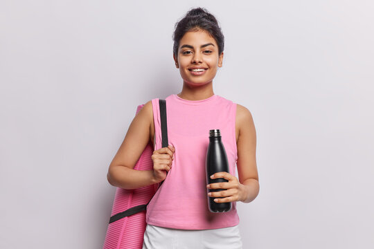 Fitness concept. Positive Iranian woman with dark hair carries rolled karemat and hold bottle of water dressed in sportswear smiles gladfully isolated over white background. Sporty lifestyle