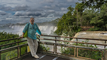 Naklejka premium The man is standing on the observation deck at the waterfalls, leaning on the railing, smiling. Behind- are the picturesque Iguazu Falls, shrouded in spray and fog. Nearby is lush tropical vegetation 