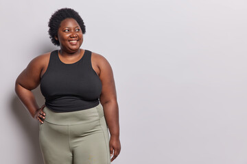 Plus size dark skinned adult woman with curly hair smiles happily keeps hand on waist wears black t...