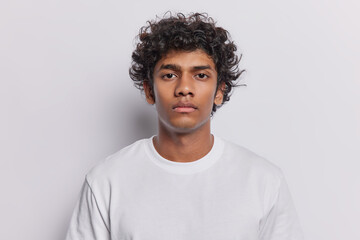 Portrait of handsome curly haired Hindu man concentrated at camera with attentive gaze poses for making photo on documents wears white clothing stands in studio. Serious boyfriend listens explanations
