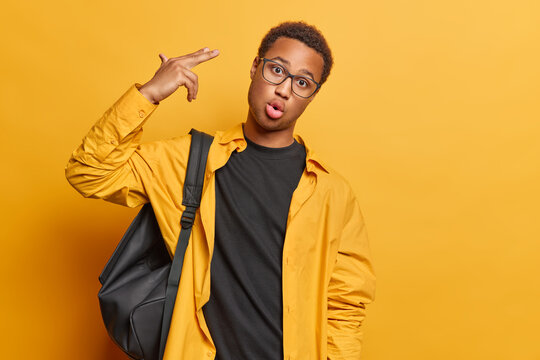 Stressed dark skinned African man makes gun gesure with fingers near temple shoots himself as has busy day at university poses in casual clothing with rucksack isolated over yellow background