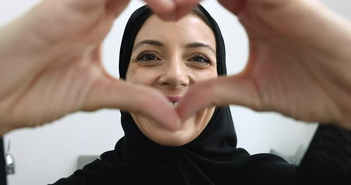 Beautiful 40s woman in traditional hijab clothes looking at camera showing heart sign with joined fingers, express love symbol, having white-toothed smile advertises dental clinic services, close up