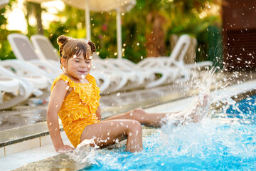 Little preschool girl playing in outdoor swimming pool by sunset. Child learning to swim in outdoor pool, splashing with water, laughing having fun. Family vacations. Healthy children sport activity