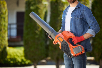 Bearded male gardener in gloves holding powerful cordless leaf blower outdoors. Crop view of...