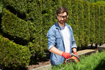 Skilled male gardener in protective glasses cutting greenery with hedge trimmer in topiary park....