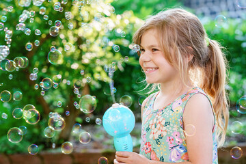 Happy little blonde preschool girl having fun with blowing soap bubble blower. Cute child playing...