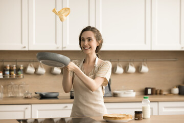 Cheerful baker girl in apron having fun in home kitchen, enjoying cooking hobby, culinary activities, preparing breakfast, frying pancakes, throwing crape over hot pan, smiling, laughing