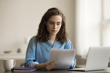 Focused young entrepreneur woman working in home office, doing paperwork accounting job at laptop, reading financial paper documents at workplace table, checking investment, insurance agreement