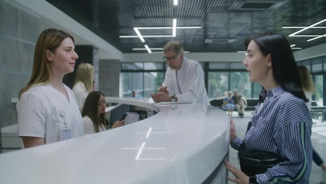 Adult woman comes to reception desk in clinic lobby area and makes appointment with doctor. Female administrator talks with patient at information counter. Medical staff work in modern medical center.