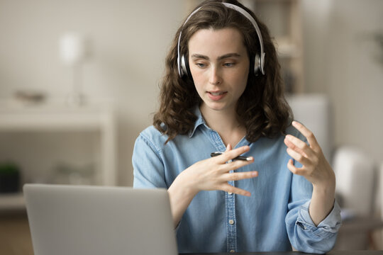 Serious gen Z student girl in headphones talking on video call at laptop, sitting at home office workplace. Online speaker streaming on Internet, giving workshop, webinar, speaking at conference