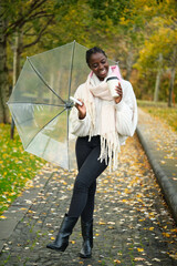 African woman with a coffee and transparent umbrella looking at camera and smiling in autumn.