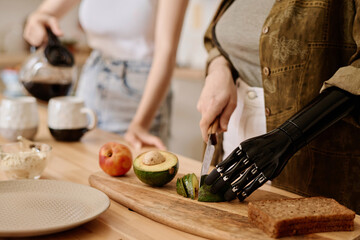 Close-up of girl with prosthetic arm cutting vegetable for breakfast while her girlfriend pouring...