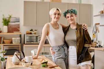 Portrait of young lesbian couple embracing and looking at camera while preparing food in the kitchen