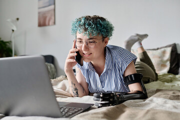 Young woman with disability having conversation on mobile phone while using laptop lying on bed in...