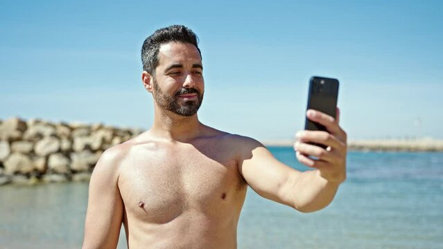 Young hispanic man tourist make selfie by smartphone standing shirtless at the beach