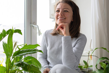 closeup portrait smilling woman sitting near house plants, flowers on window. happy female in cozy...