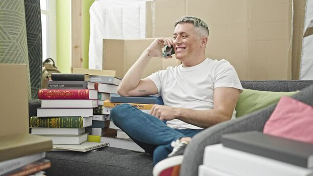 Young caucasian man talking on smartphone sitting on sofa at new home