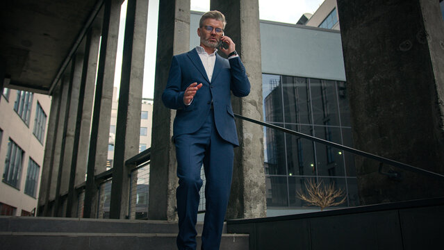 Walking outdoors going old mature Caucasian business man employer talk smartphone negotiate speaking cellphone walk in city near office company building talking mobile phone call senior businessman