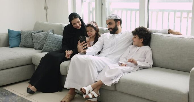 Family with little children in traditional Islamic clothes use smartphone at home seated on couch. Virtual meeting event with relatives, using mobile application for fun, make selfie pictures on cell