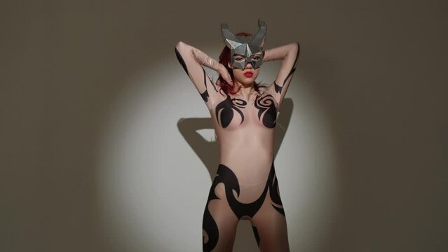 A devil girl with horns on her head in a sexy tight costume dances beautifully in the studio on a gray background.