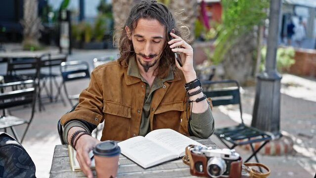Young hispanic man tourist talking on smartphone sitting on table taking notes at coffee shop terrace
