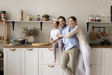 Cheerful mom, dad, little toddler kid posing for home portrait in kitchen, leaning on new furniture, standing and hugging in modern interior with utensil, looking at camera, smiling, laughing